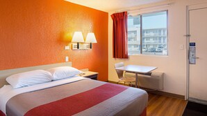A hotel room with a bed, a desk, a chair, a window with a view of buildings, and a door.