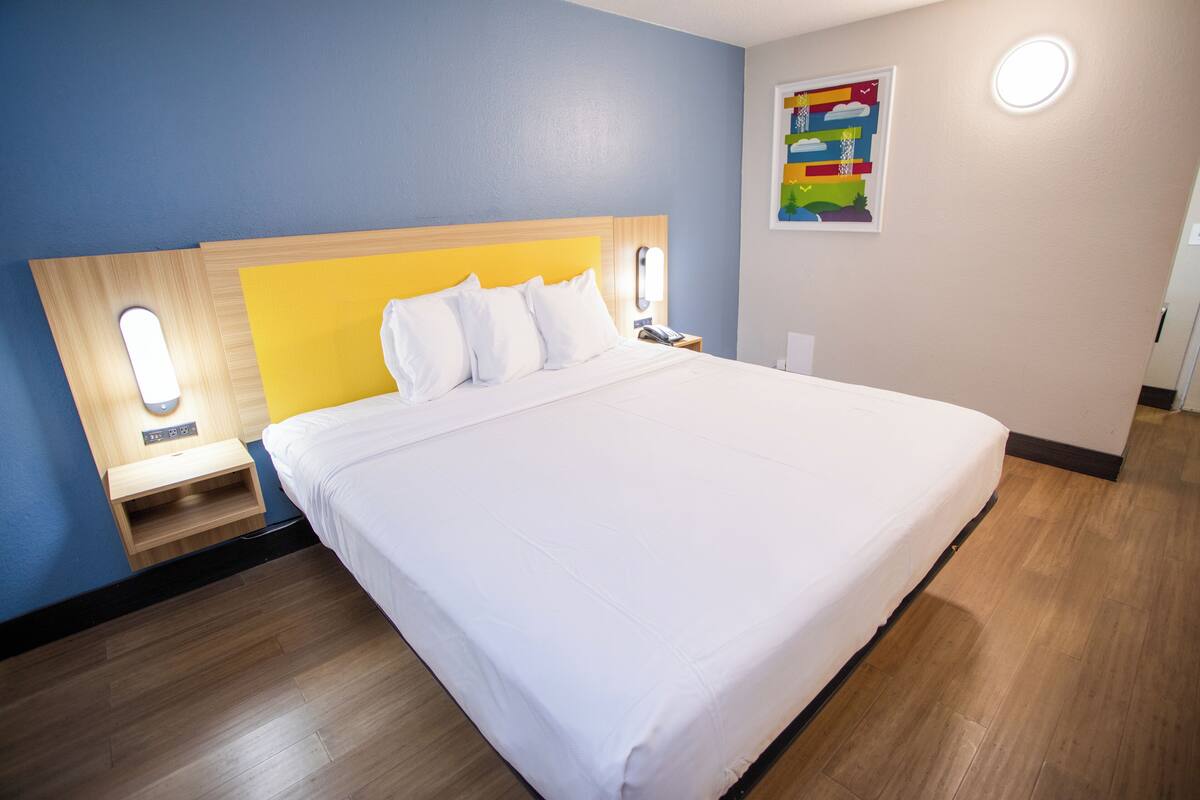 A hotel room with a large bed, wooden headboard, bedside tables with lamps, and a colorful artwork on the wall.