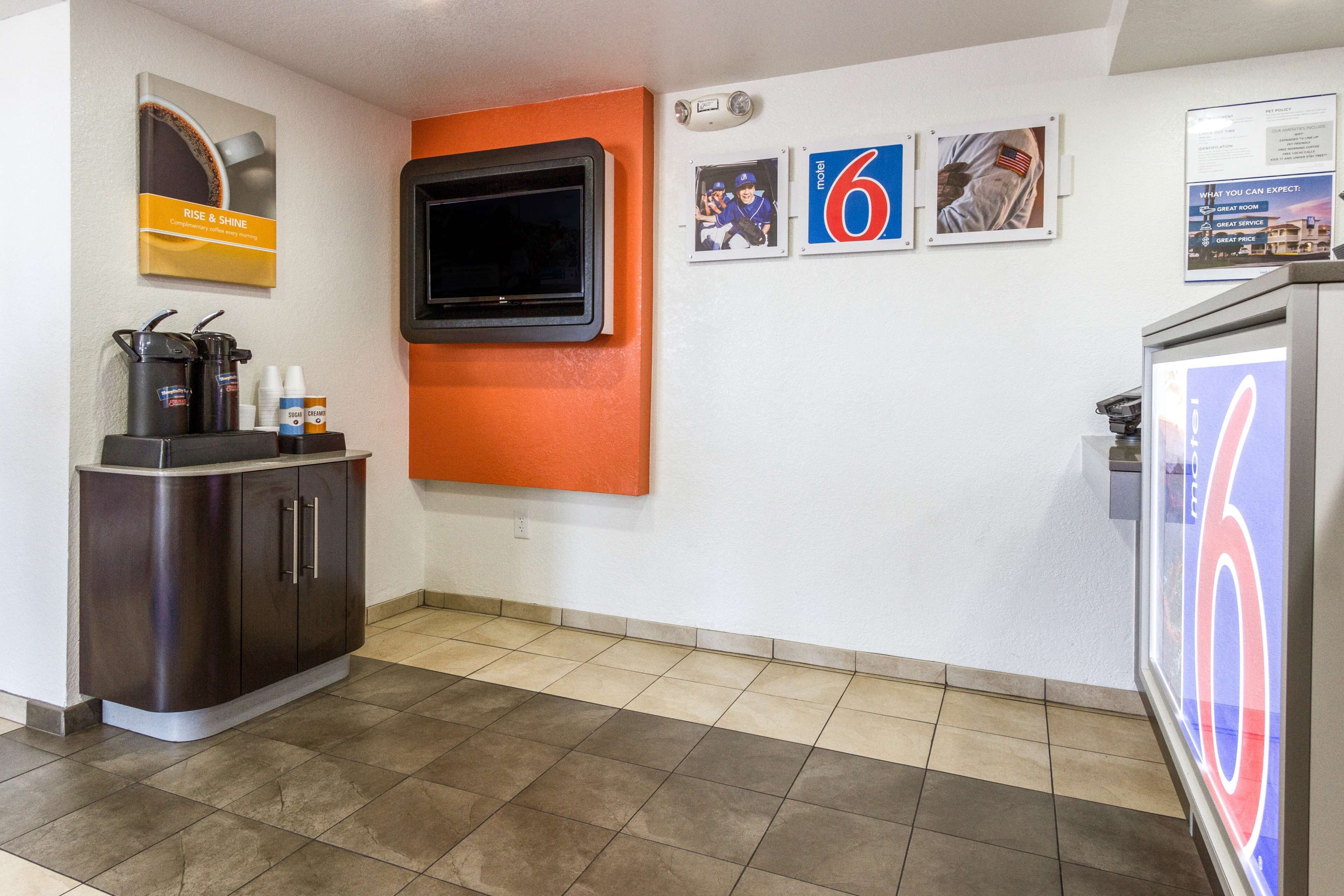 A hotel lobby with a coffee station, a TV mounted on an orange wall, and a refrigerator with a '6' sign.