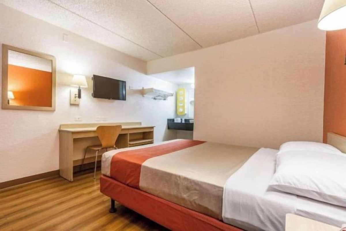 Una habitación de hotel con cama, escritorio, silla, televisor y espejo.