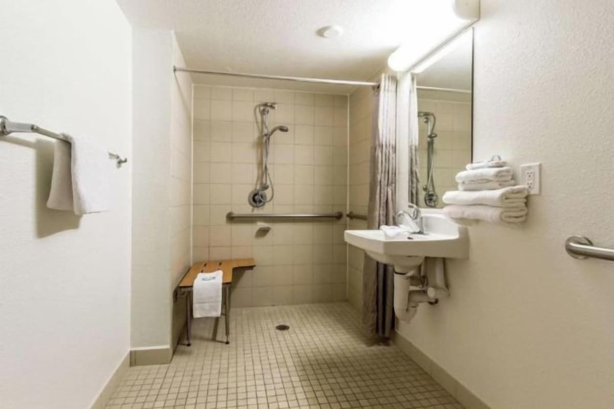 A small, clean bathroom with a shower, a sink, and a mirror. There is a bench and a towel rack.