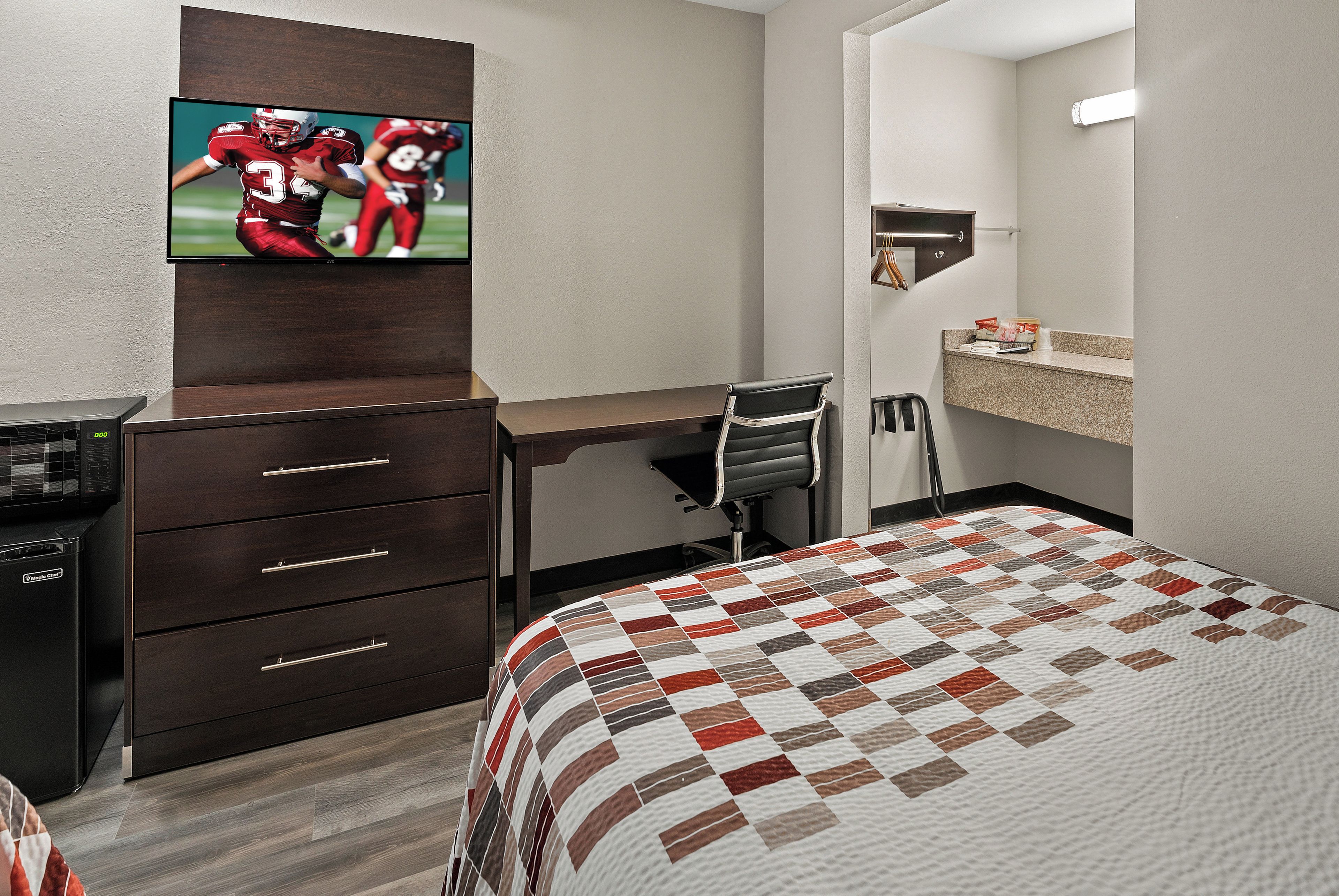 A hotel room with a bed, a desk, a chair, a TV mounted on a wall, and a minibar.