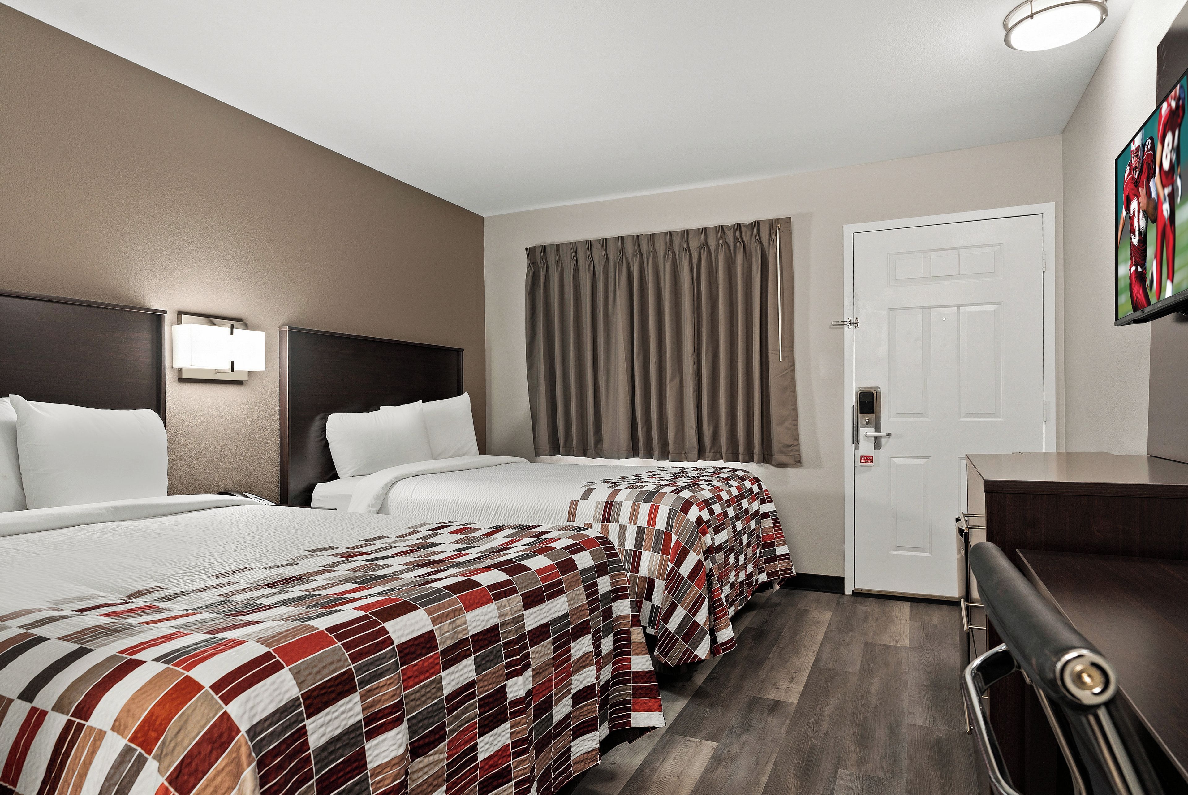 A hotel room with two beds, a desk, a door, and a window with curtains.