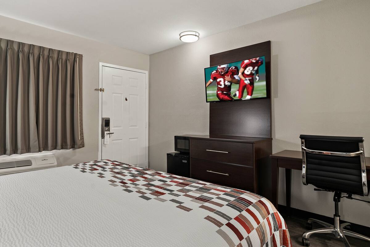 A hotel room with a bed, a TV mounted on the wall, a desk with a chair, and a door.