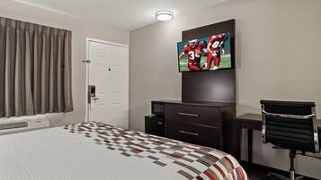A hotel room with a bed, a television mounted on the wall, a desk with a chair, and a window with curtains.