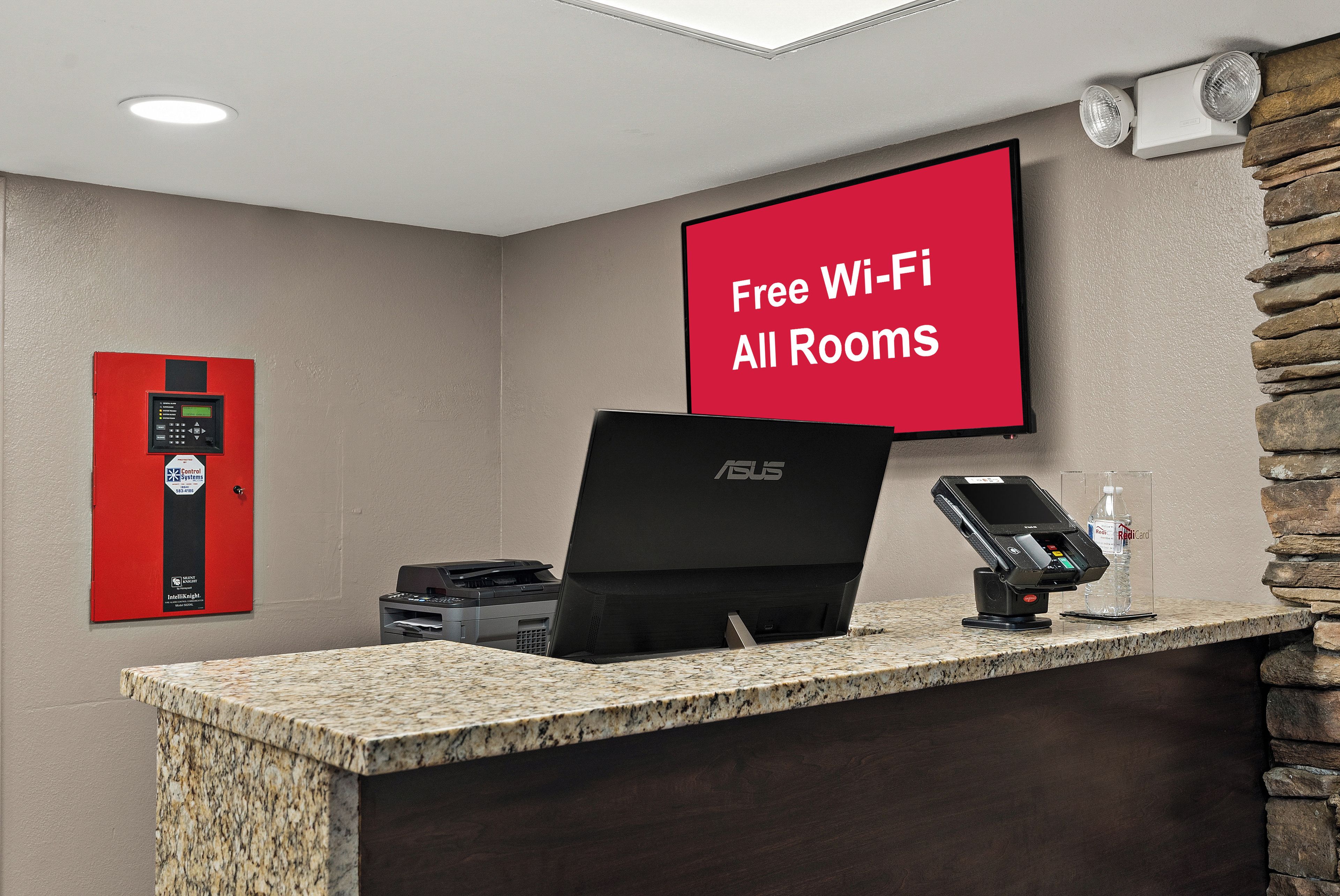 Et hotellets receptionområde med et rødt skilt, der angiver gratis Wi-Fi på alle værelser, et kontrolpanel, en bærbar computer og en betalingsterminal.
