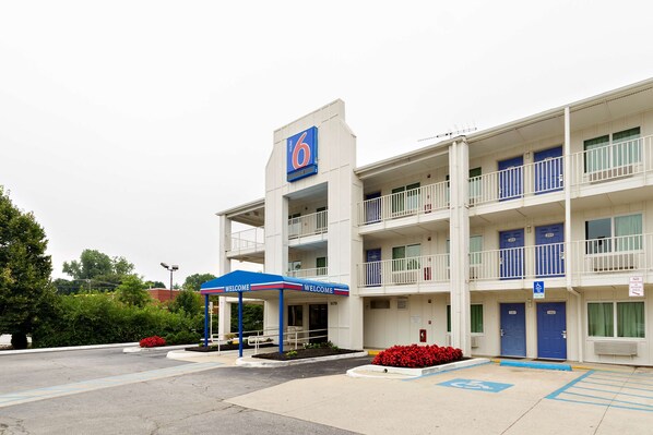 Un motel de varios pisos con un llamativo letrero con el número '6', un acceso cubierto y un estacionamiento con accesibilidad para personas con discapacidad.