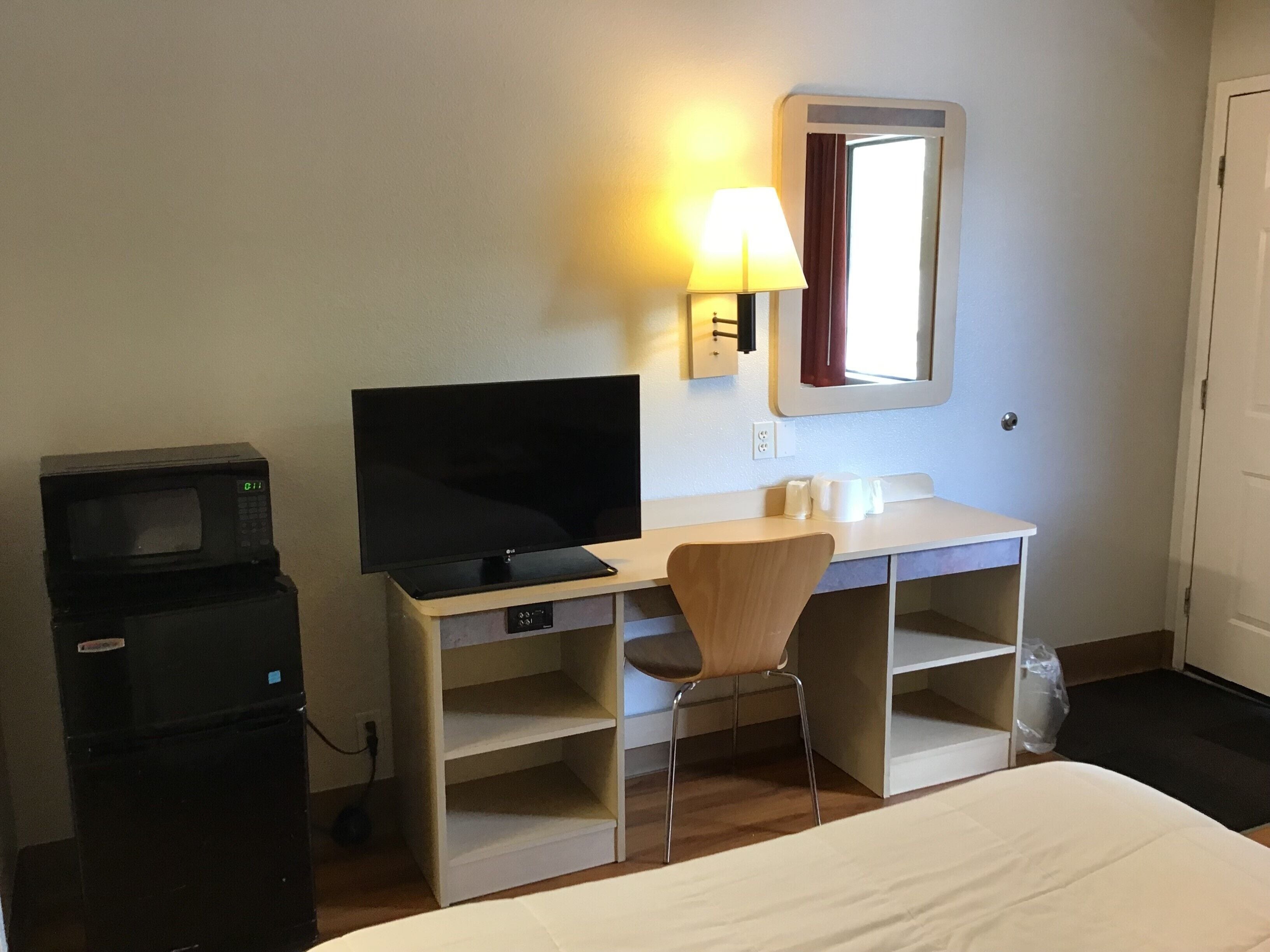 Habitación de hotel con cama, escritorio con televisor, silla, microondas y refrigerador pequeño.