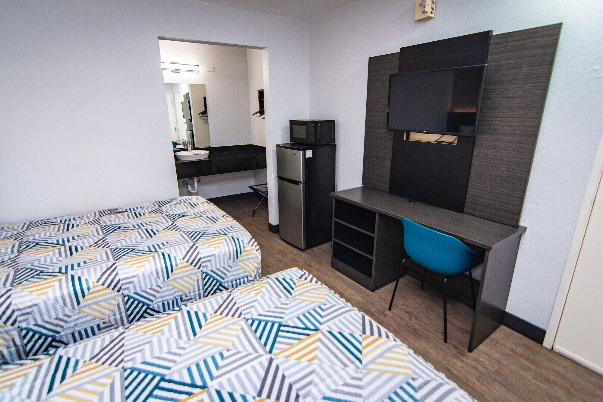 A compact hotel room with two beds, a kitchenette, a small desk, and a TV.
