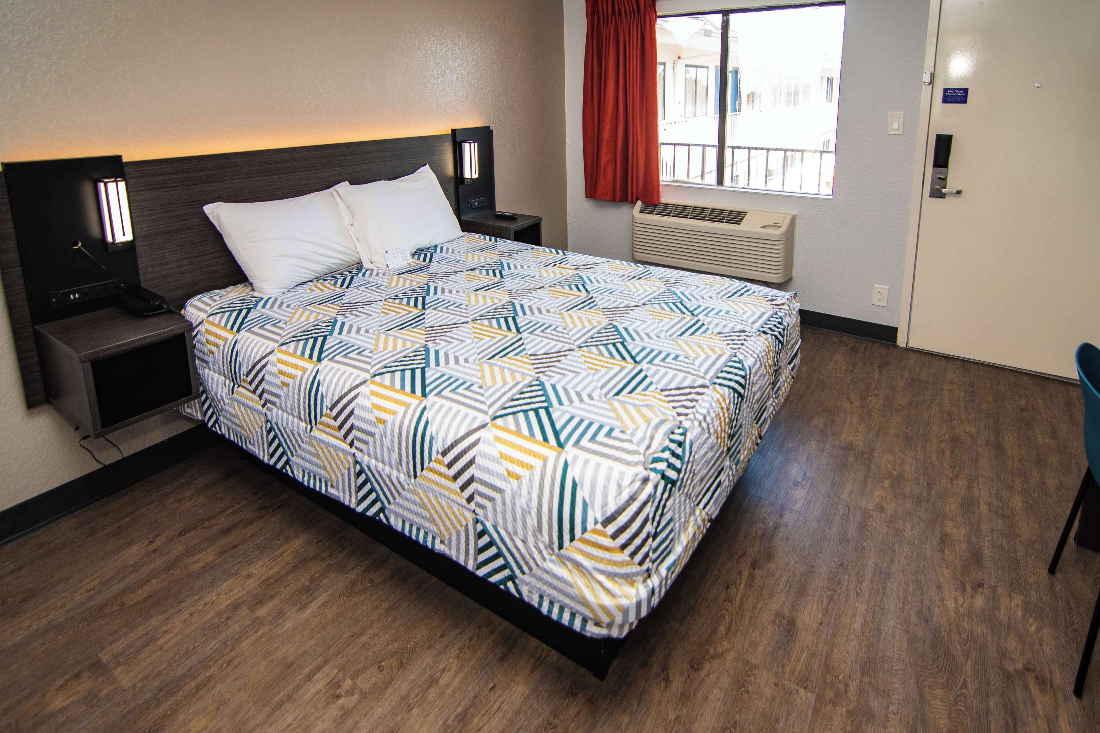 A hotel room with a bed, nightstands, a window with curtains, an air conditioning unit, and a door.