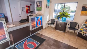 A motel reception area with a desk, chairs, a plant, and a window with a view of the parking lot.