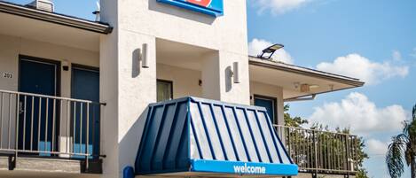 A motel with a prominent '6' sign, a 'welcome' sign, and a balcony with a railing.