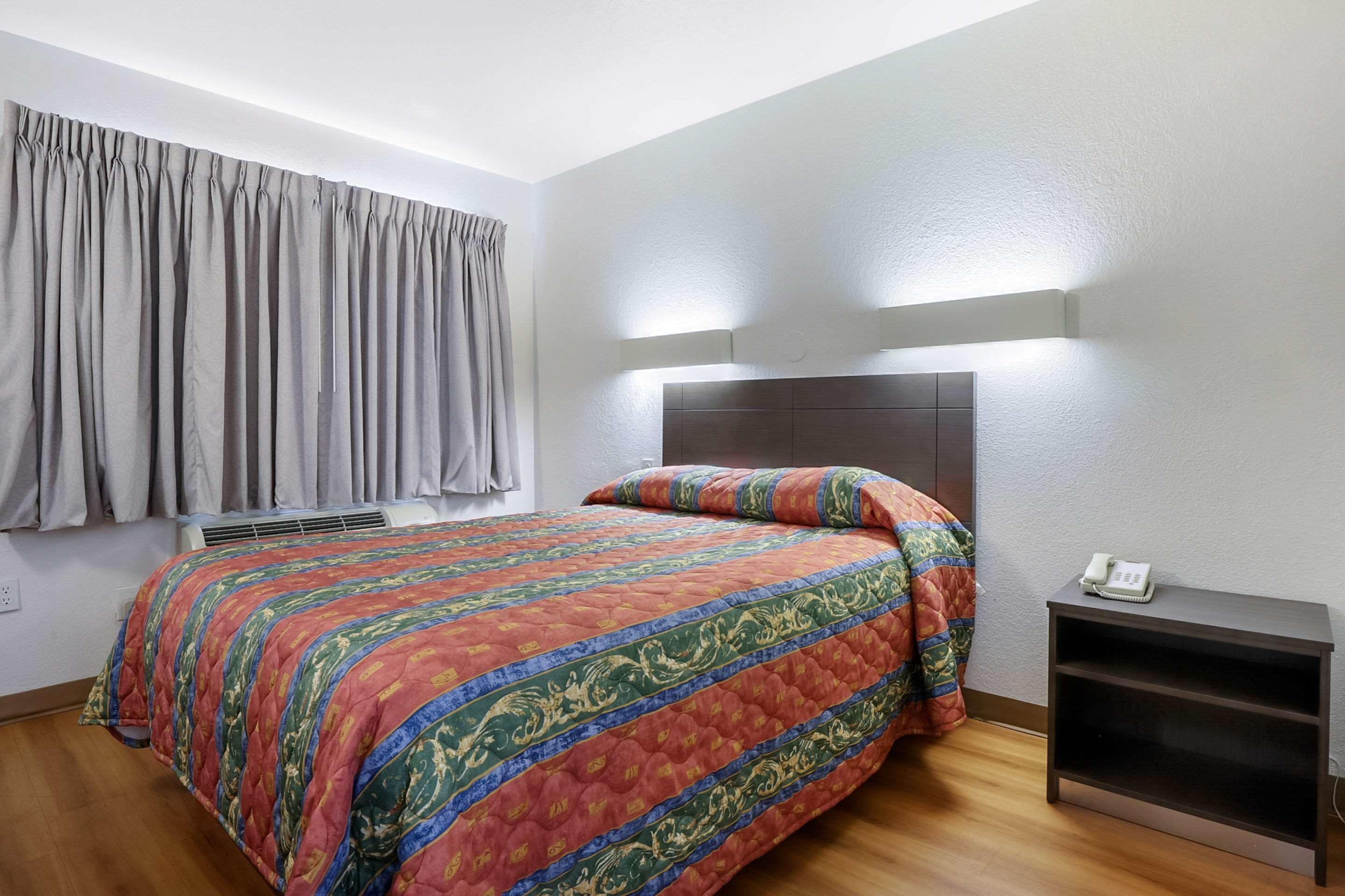 Een slaapkamer met een bed, grijze gordijnen, een nachtkastje met een telefoon en een wandlamp.