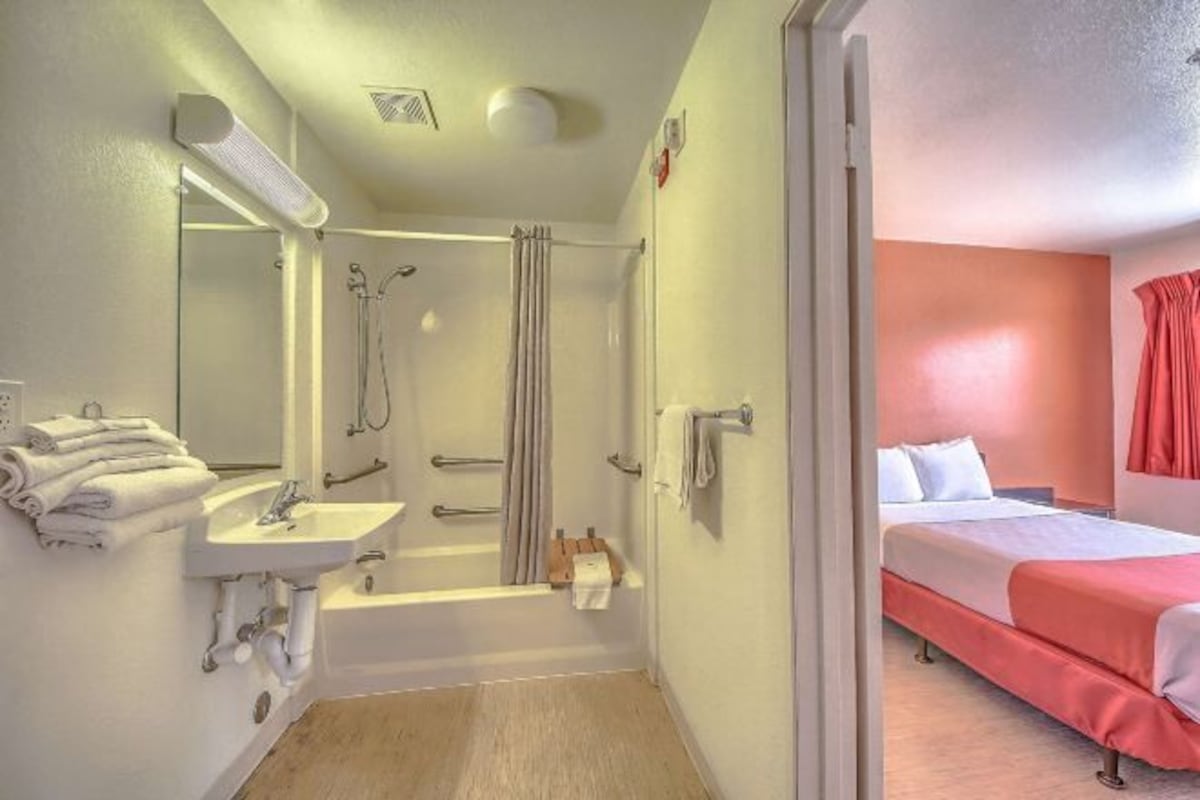 Un baño con ducha, lavabo e inodoro. En la habitación contigua, hay una cama con una manta roja.