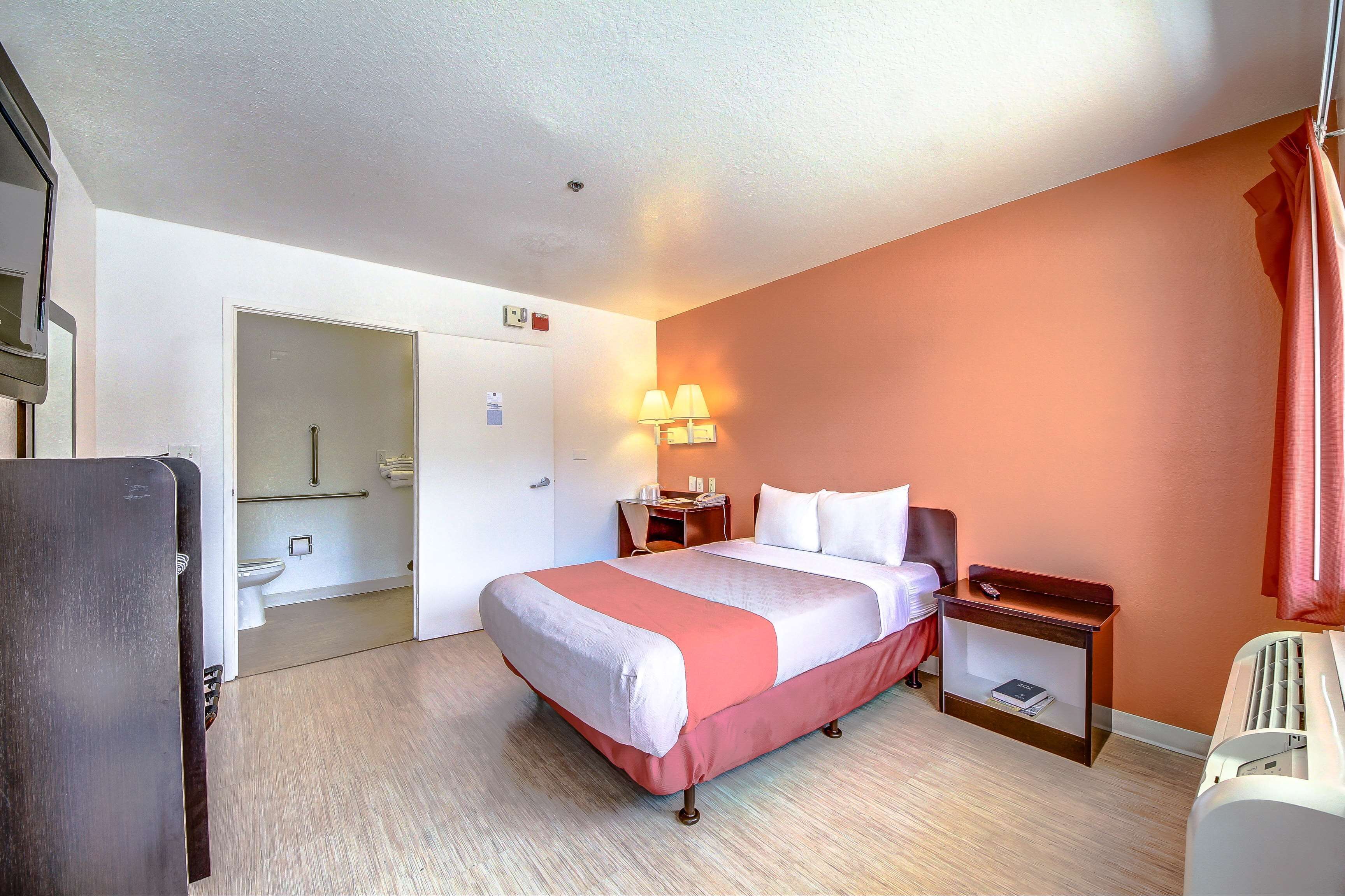 Habitación de hotel con cama, mesita de noche, lámpara y vista al baño.