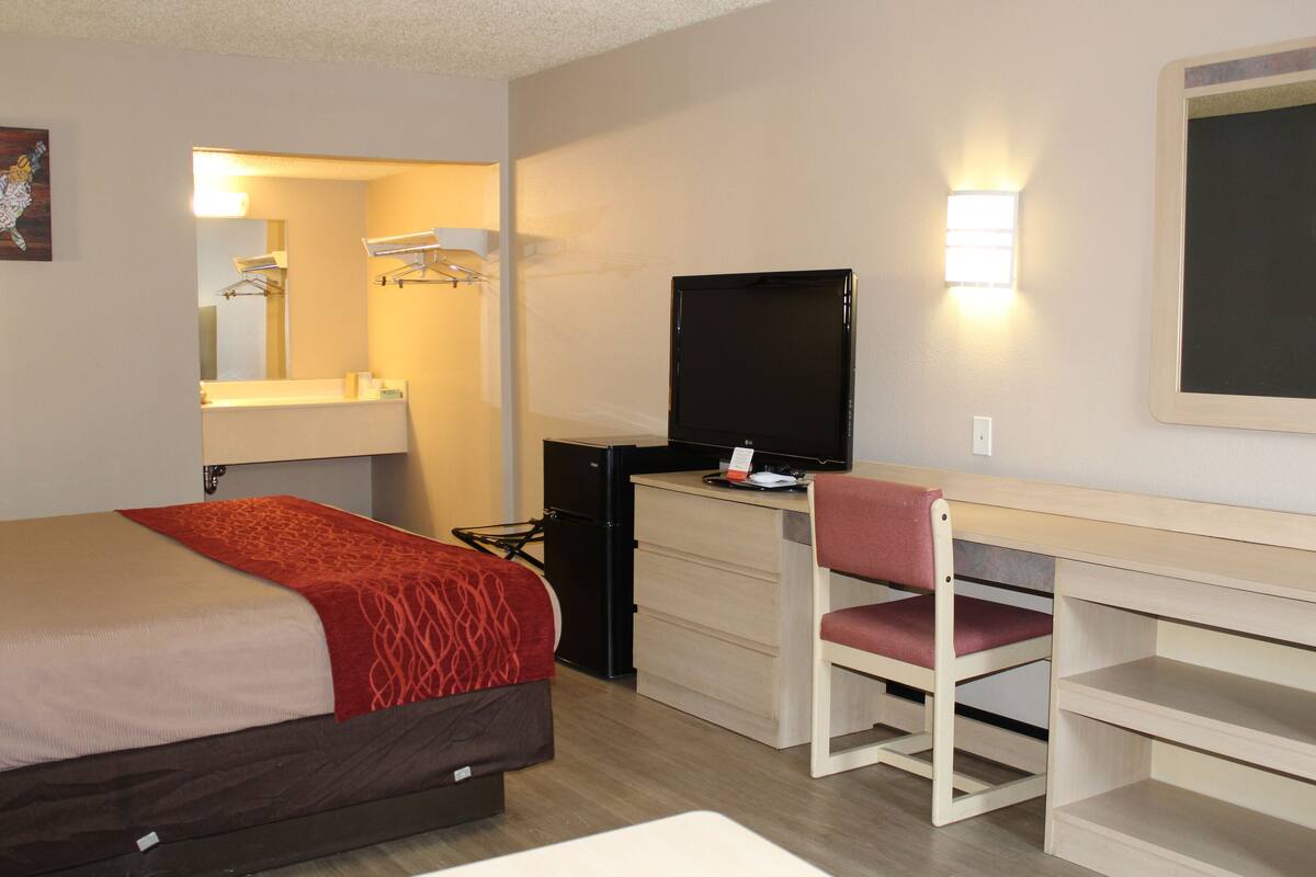 A hotel room with a bed, a desk with a chair, a television, a mirror, and a shelf.