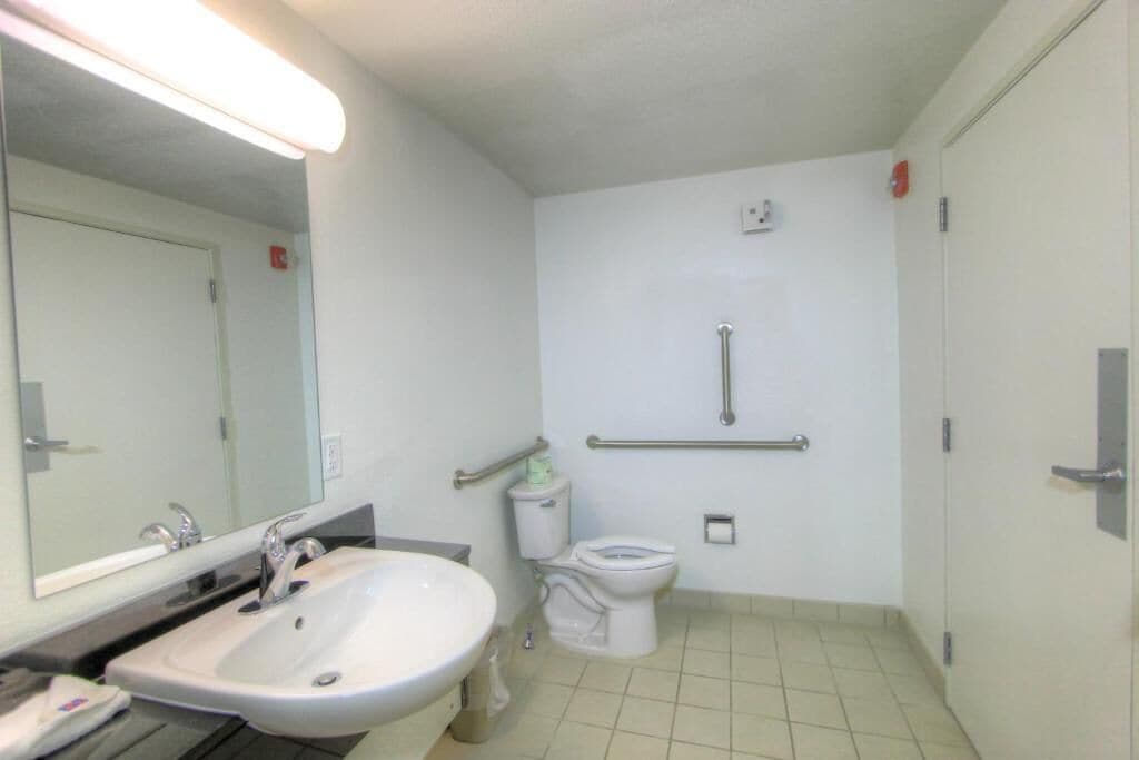 A small, clean bathroom with a sink, mirror, toilet, and grab bars.