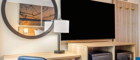 A hotel room with a desk, chair, television, microwave, and a mirror.