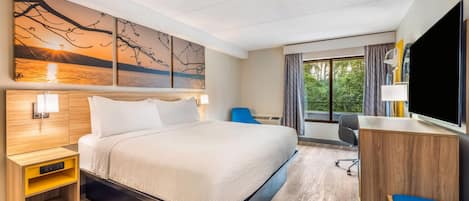 A hotel room with a bed, bedside table, desk, chair, and a large window.