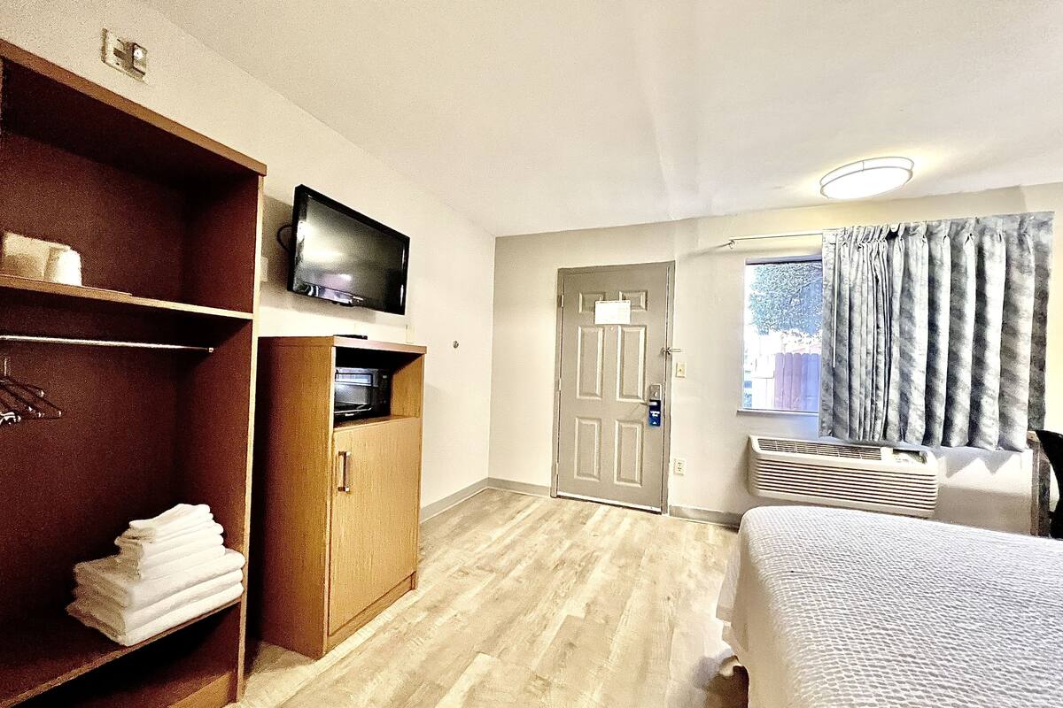 Ein Hotelzimmer mit einem Bett, einem Fernseher auf einem Regal, einer Tür und einem Fenster mit Vorhängen.