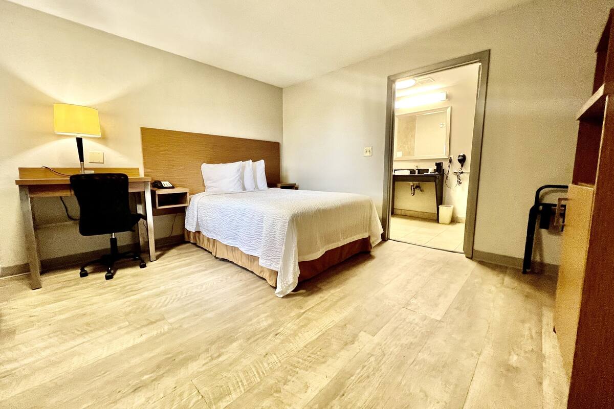Ein Hotelzimmer mit Bett, Schreibtisch mit Lampe, Stuhl und einem durchgegangenen Tür sichtbaren Badezimmer.