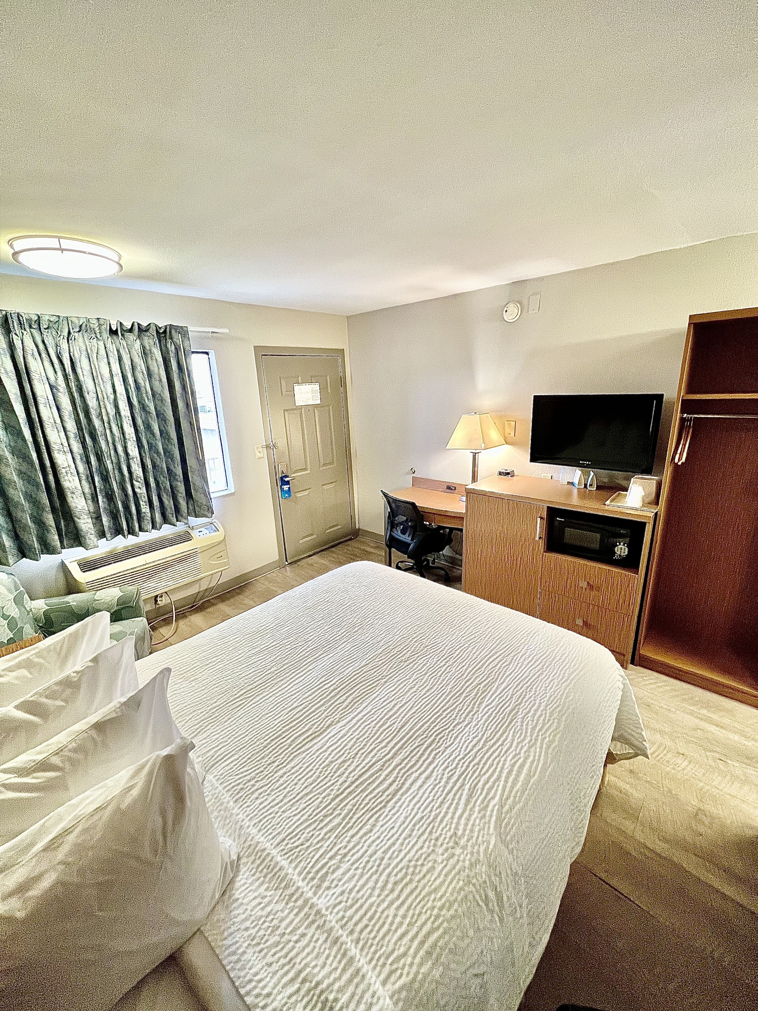 Quarto de hotel com cama, escrivaninha, computador, televisão e janela com cortinas.