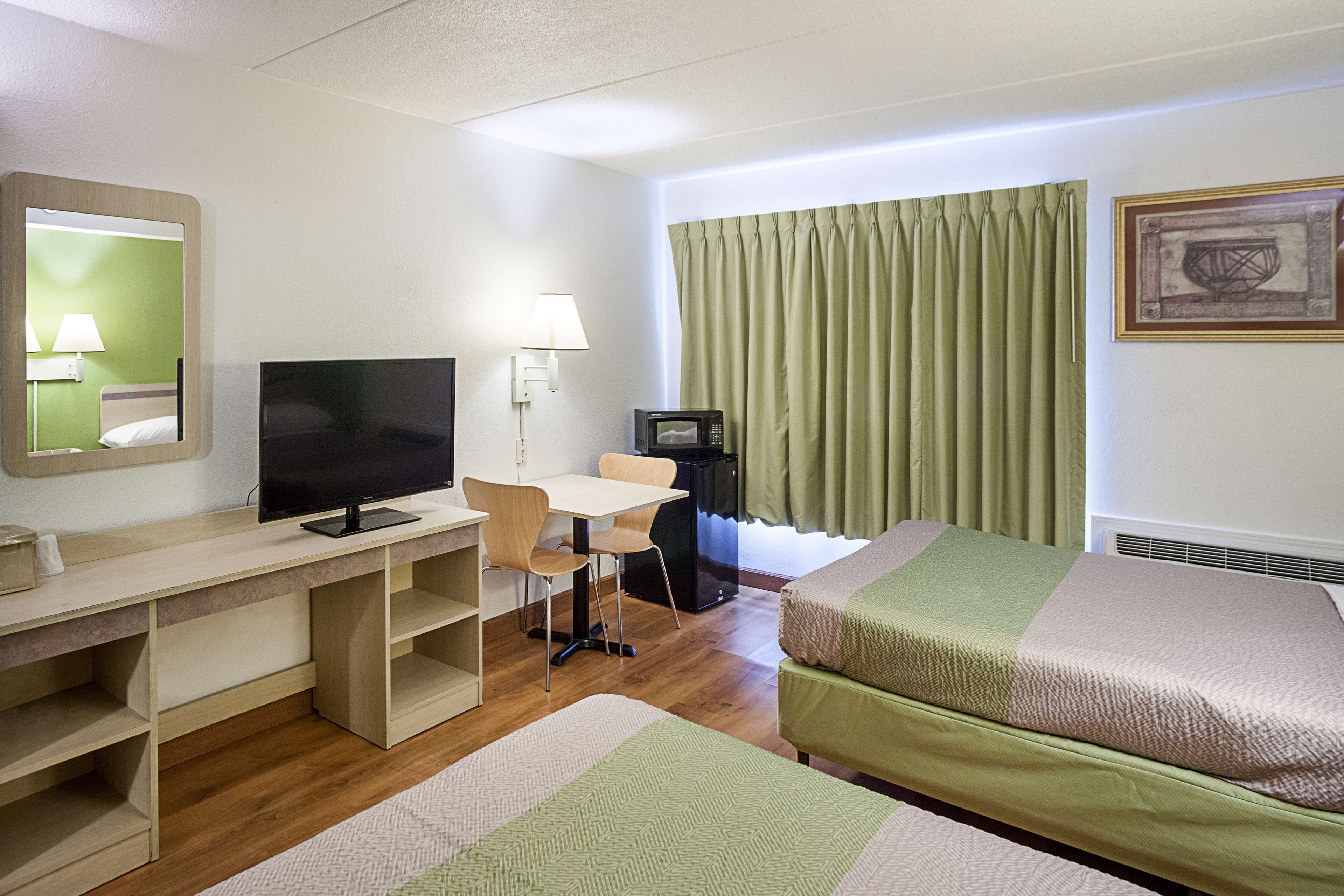 Una habitación de hotel con una cama, un escritorio con computadora, televisor, lámpara y ventana con cortinas.