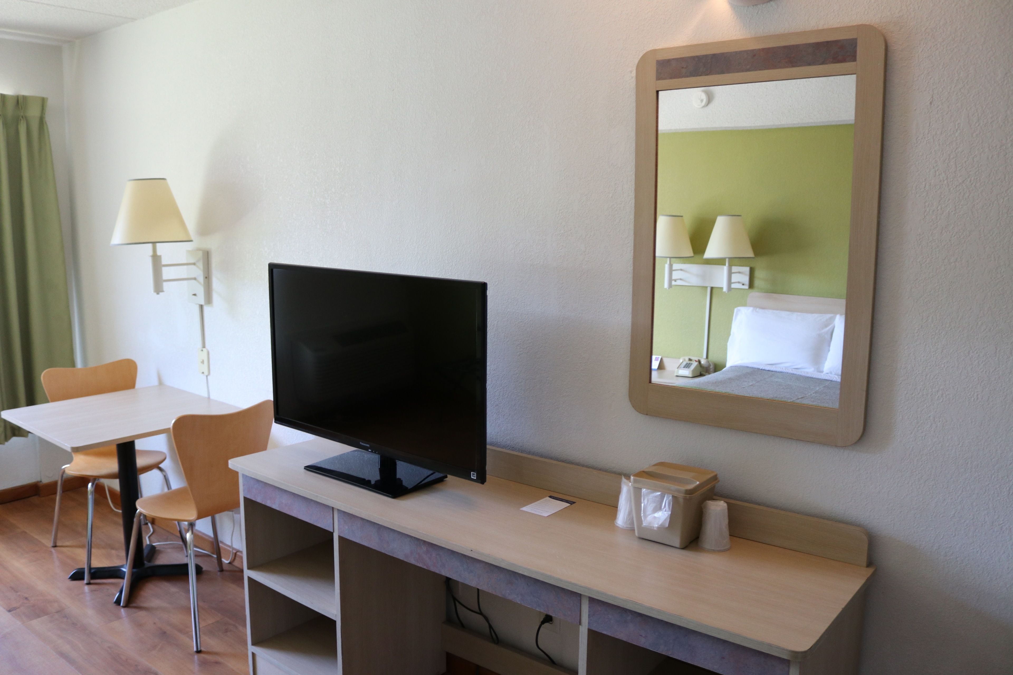 Camera d'albergo con una scrivania, una TV a schermo piatto, uno specchio, una lampada e una tenda verde.