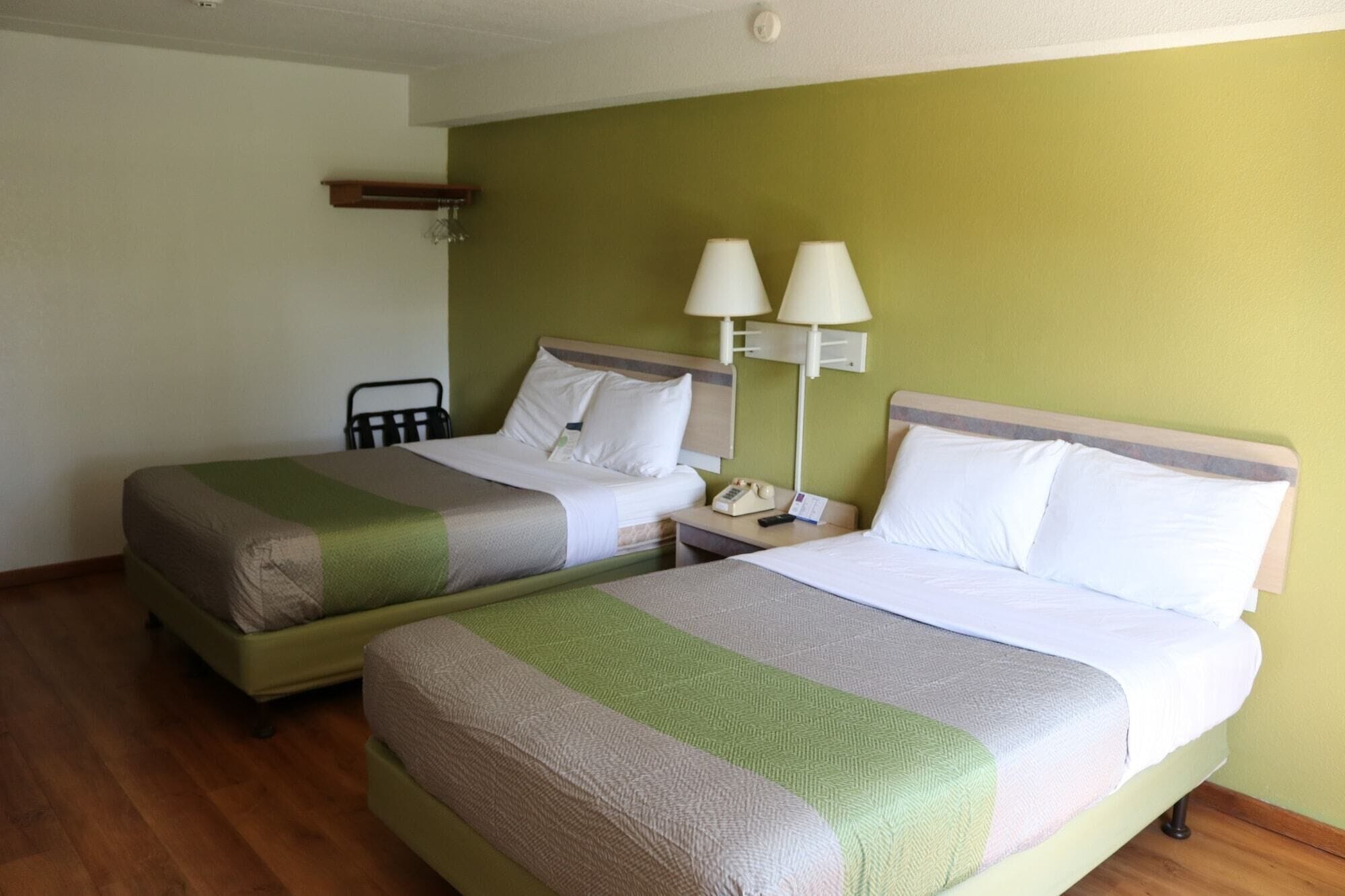 Two single beds with green and white bedding, a nightstand with a lamp, and a wooden shelf on the wall.