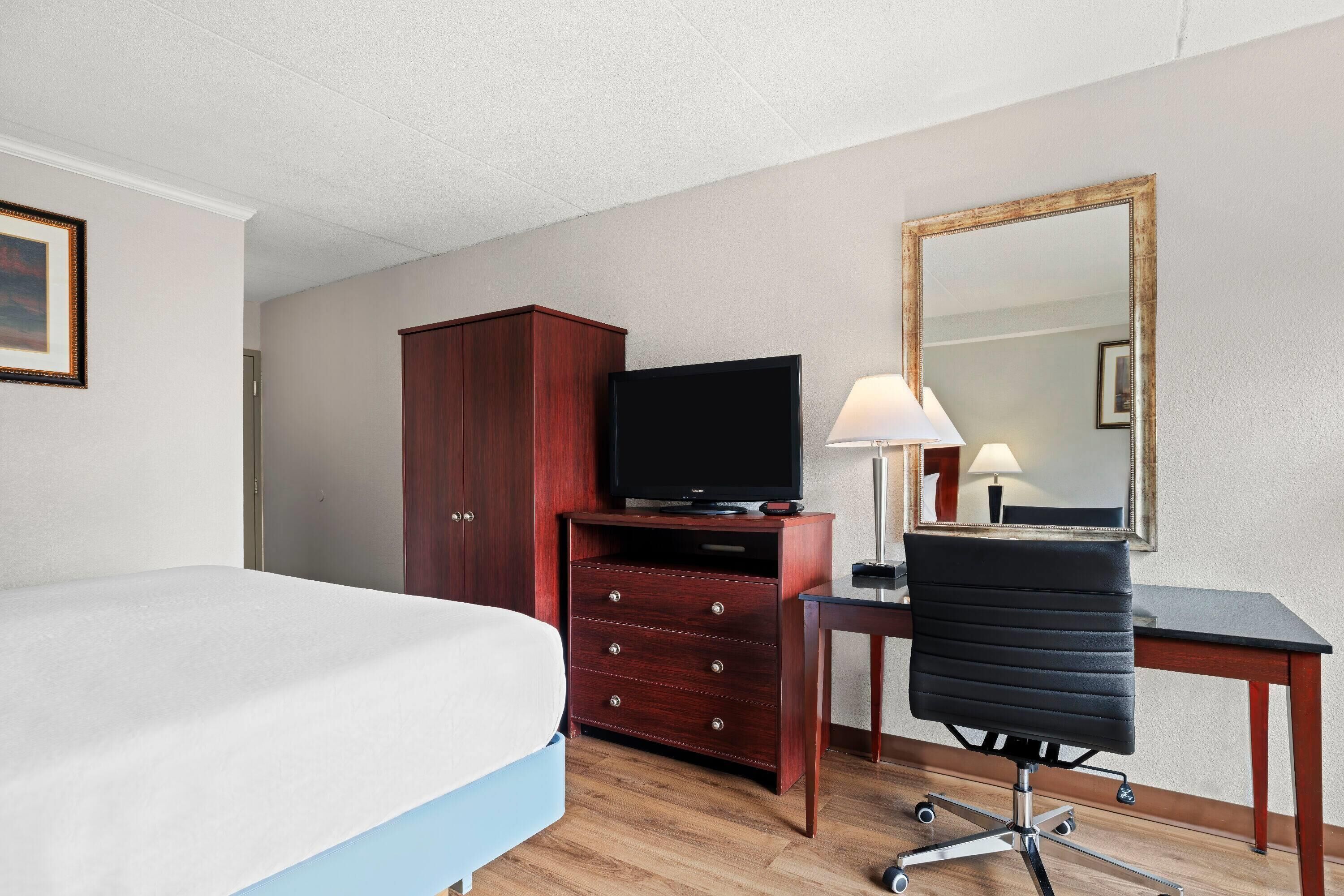 A hotel room with a bed, a wooden dresser with a television, a desk with a chair, and a large mirror on the wall.