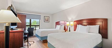 Quarto de hotel com duas camas, uma escrivaninha com computador, uma cadeira e um abajur.