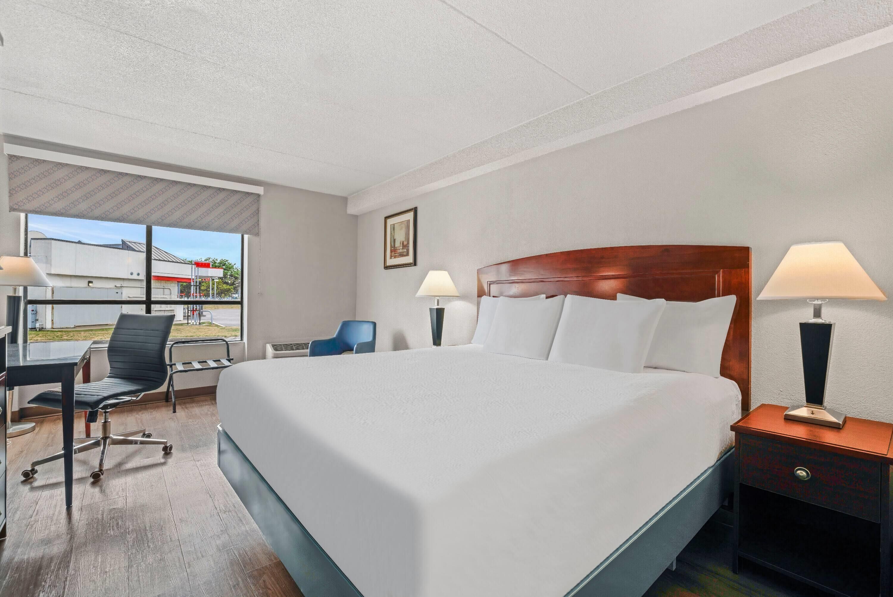 Une chambre d’hôtel avec un grand lit, un bureau avec une chaise, deux lampes de chevet et une fenêtre donnant sur un immeuble.