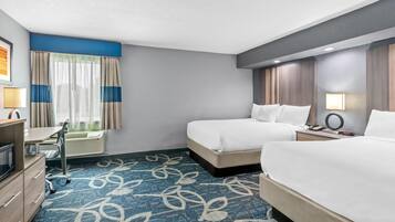 Habitació d'hotel amb dos llits, un escriptori, una cadira, un microones, una làmpada i una finestra amb cortines.