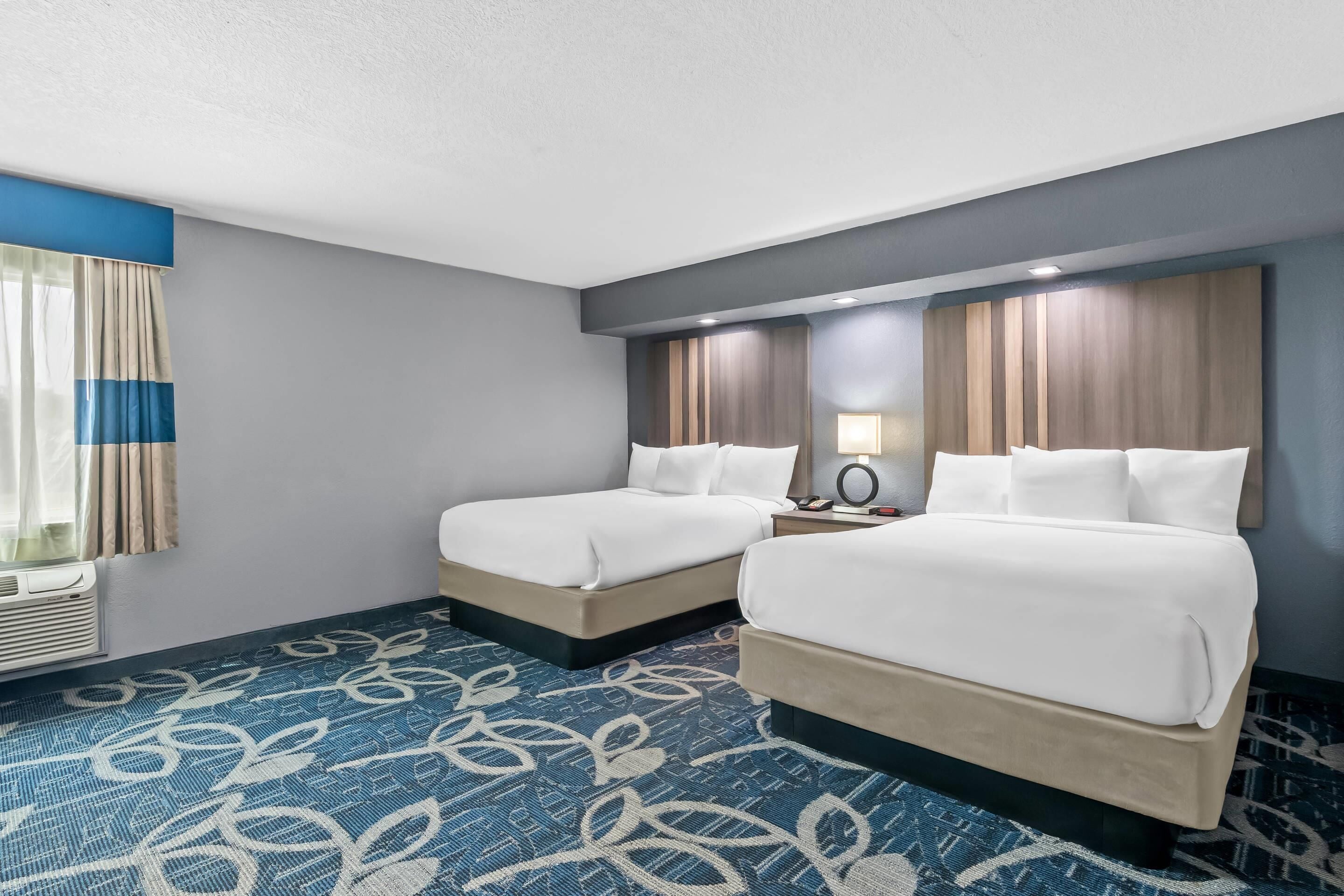 A hotel room with two beds, a patterned carpet, a window with curtains, and a wall-mounted air conditioning unit.