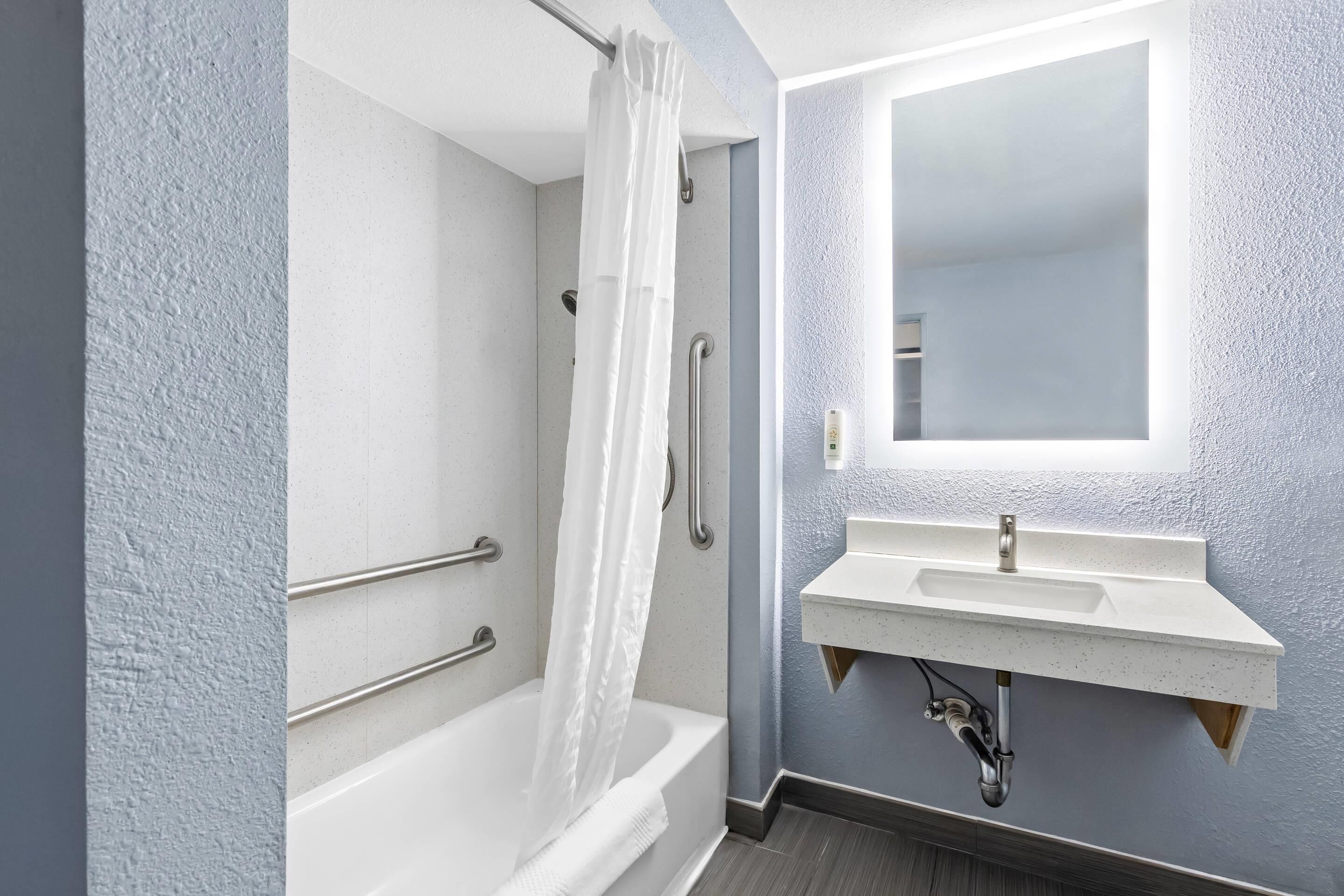 Un baño con bañera, ducha con cortina, lavabo con espejo y barra de apoyo.
