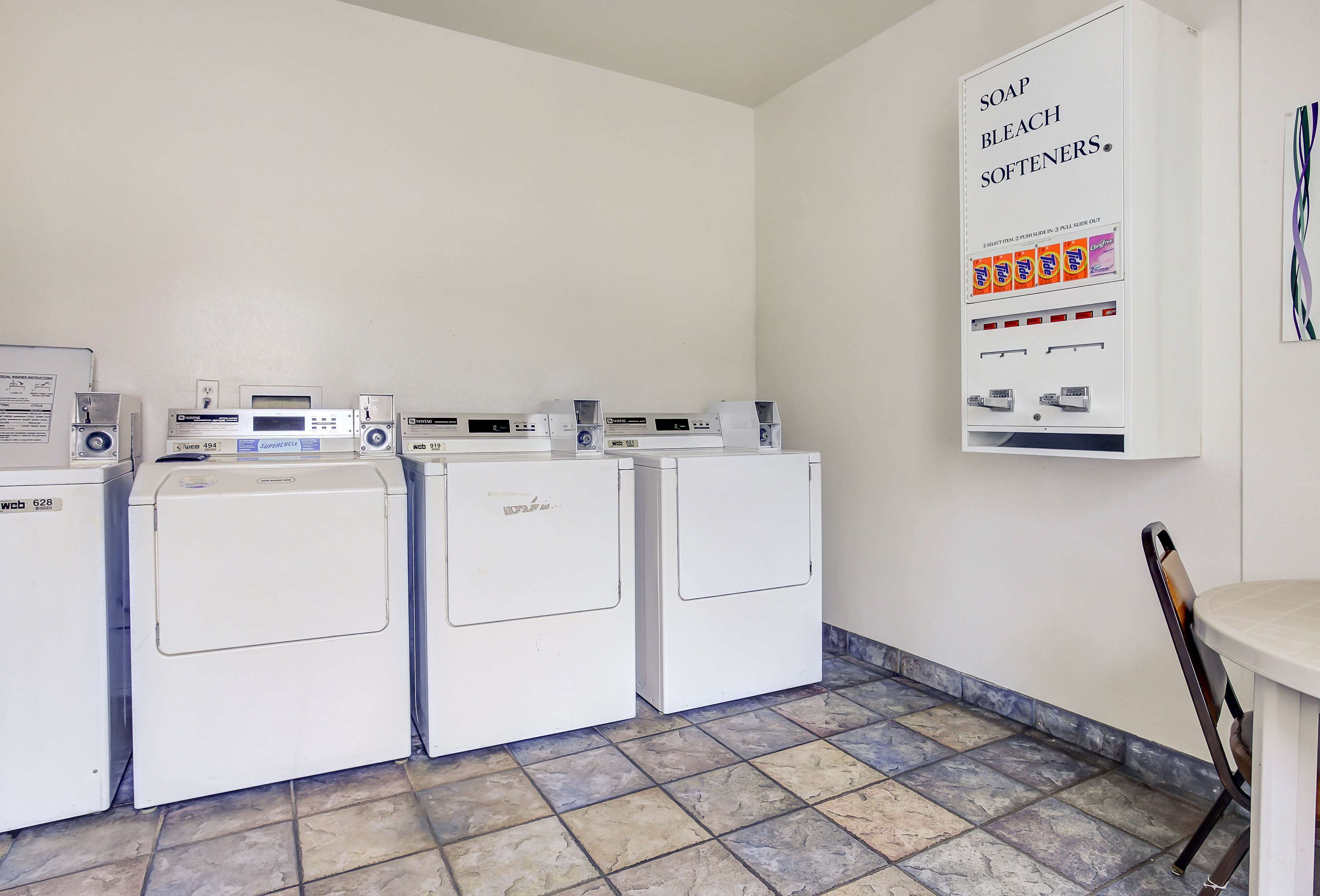 A laundry area with four white washers and dryers, a wall-mounted soap and bleach dispenser, and a small table with a chair.