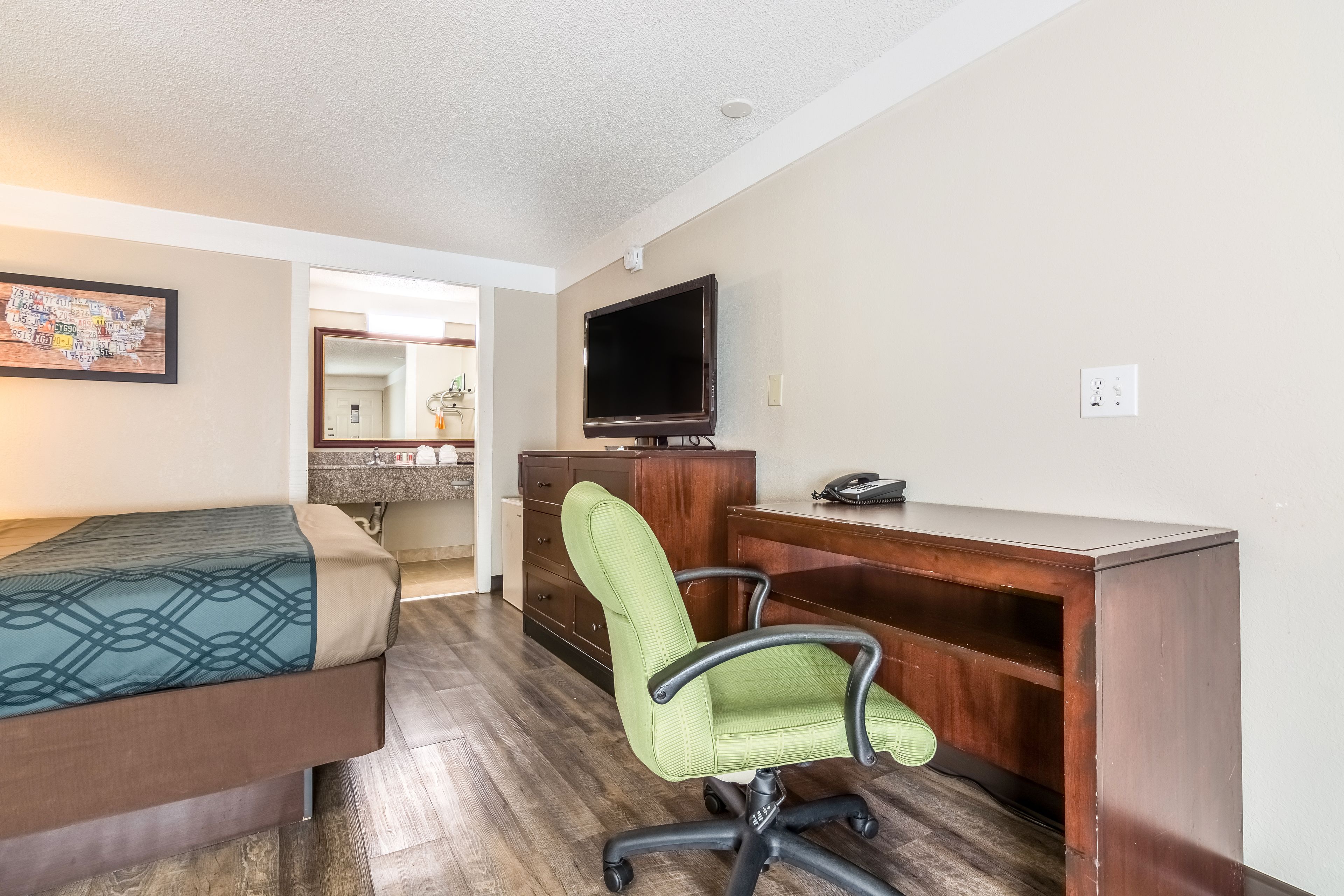 A hotel room with a bed, a desk with a chair, a television, and a bathroom visible through an open door.