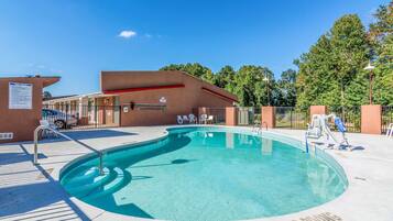 A swimming pool with a sunbathing area, a covered seating area, and a building with a red roof.