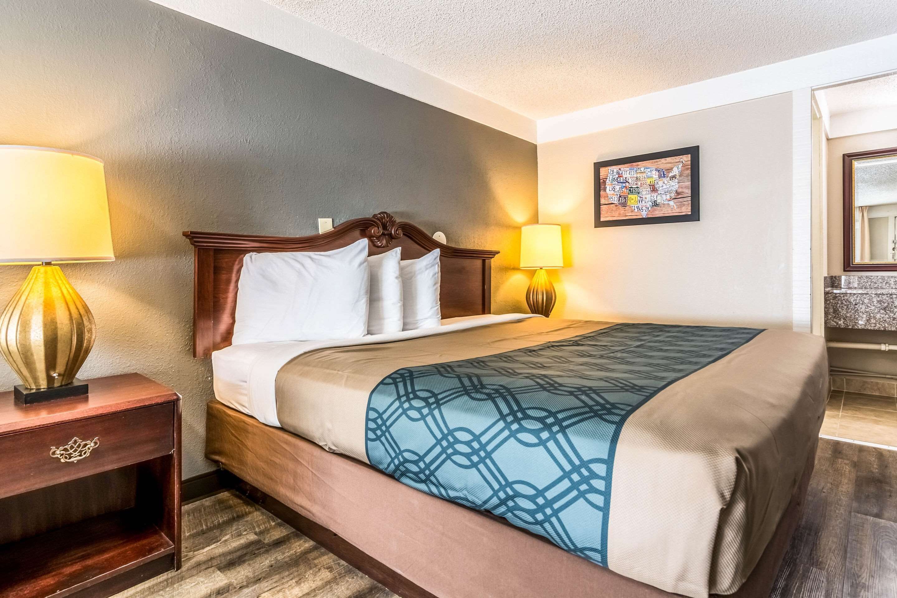 A hotel room with a bed, bedside lamps, a nightstand, a framed artwork on the wall, and a bathroom visible in the corner.