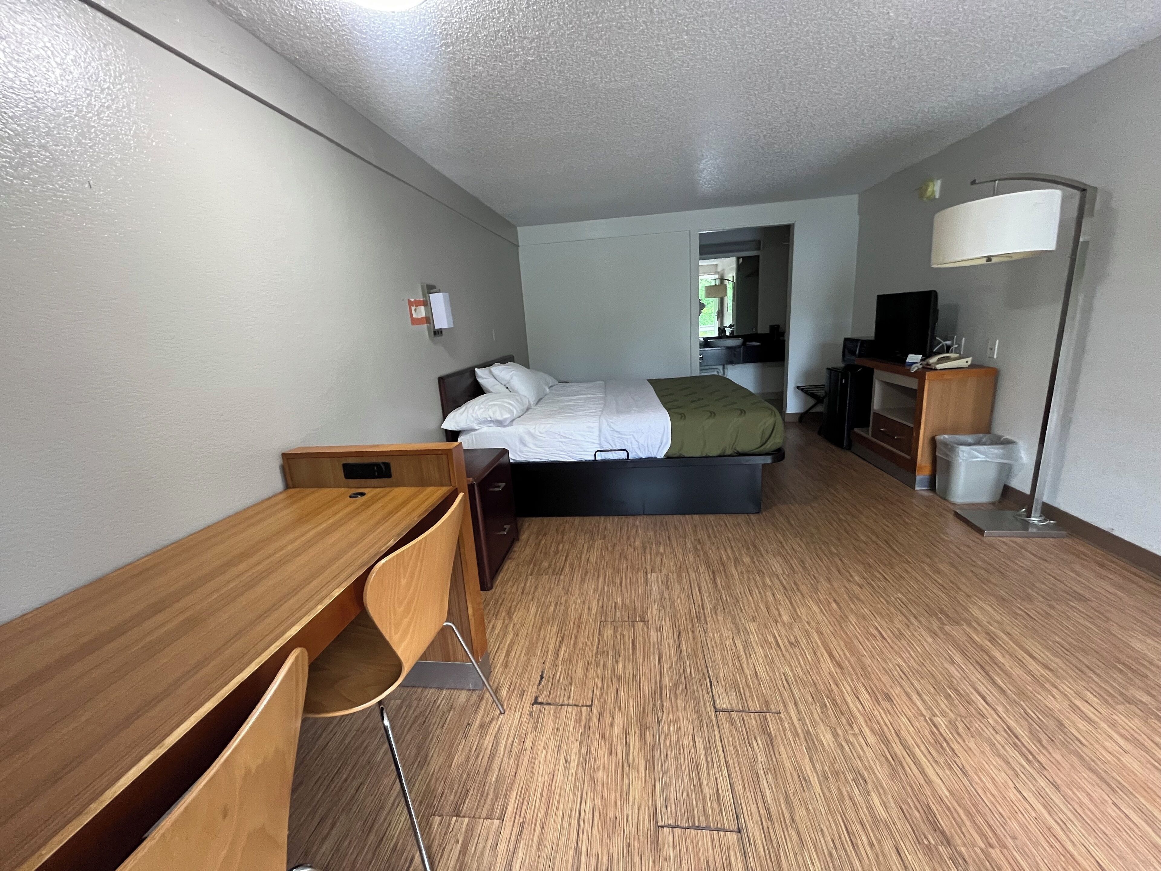 Quarto de hotel com escrivaninha de madeira, cama com roupa de cama branca, manta verde, televisão, luminária e um cesto de lixo.