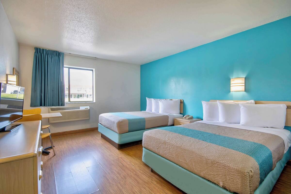 A hotel room with two beds, a desk, a chair, a window with curtains, and a wall-mounted light fixture.