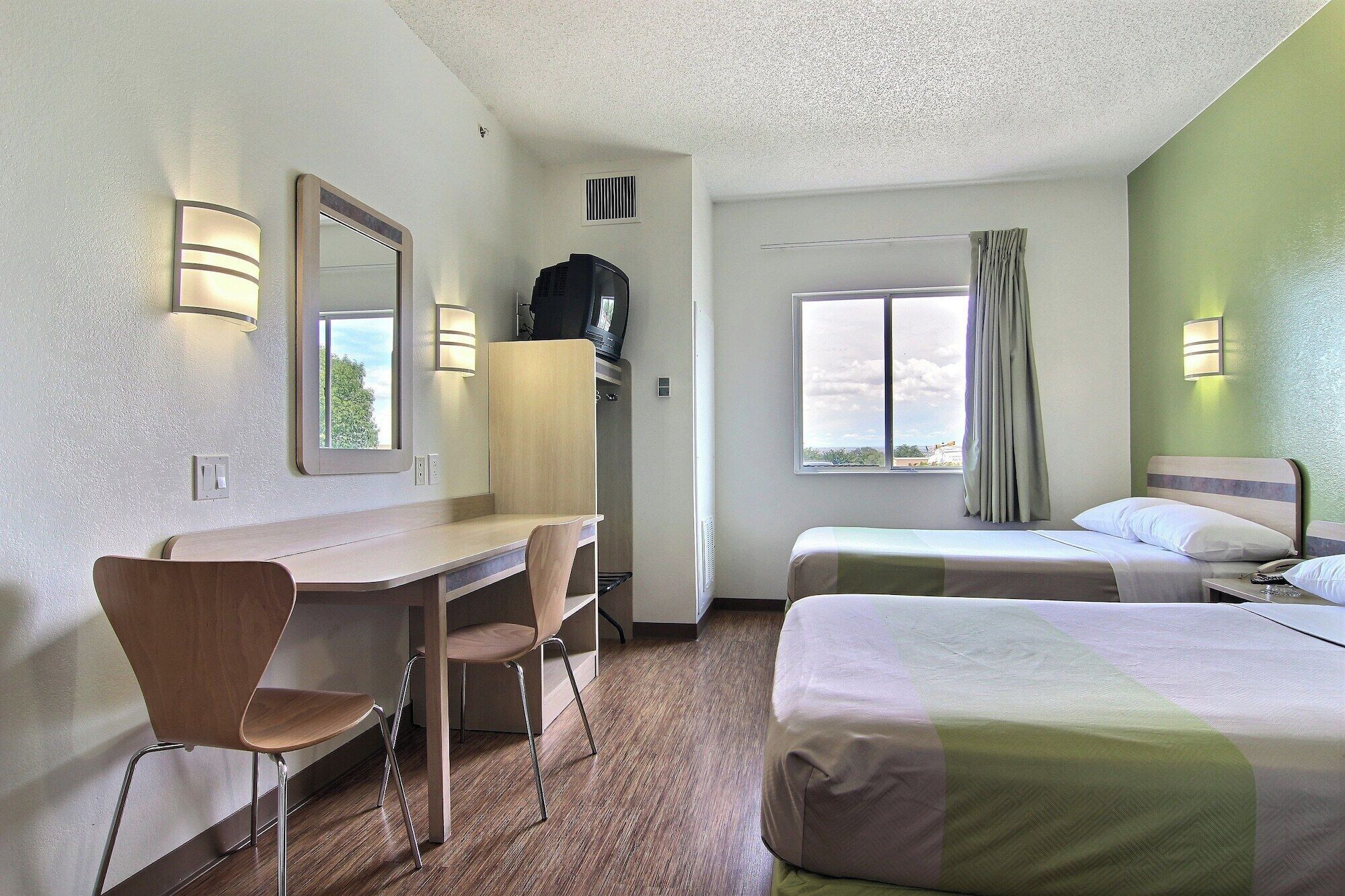 Ett hotellrum med ett skrivbord, två sängar, en stol, en spegel och ett fönster med gardiner.