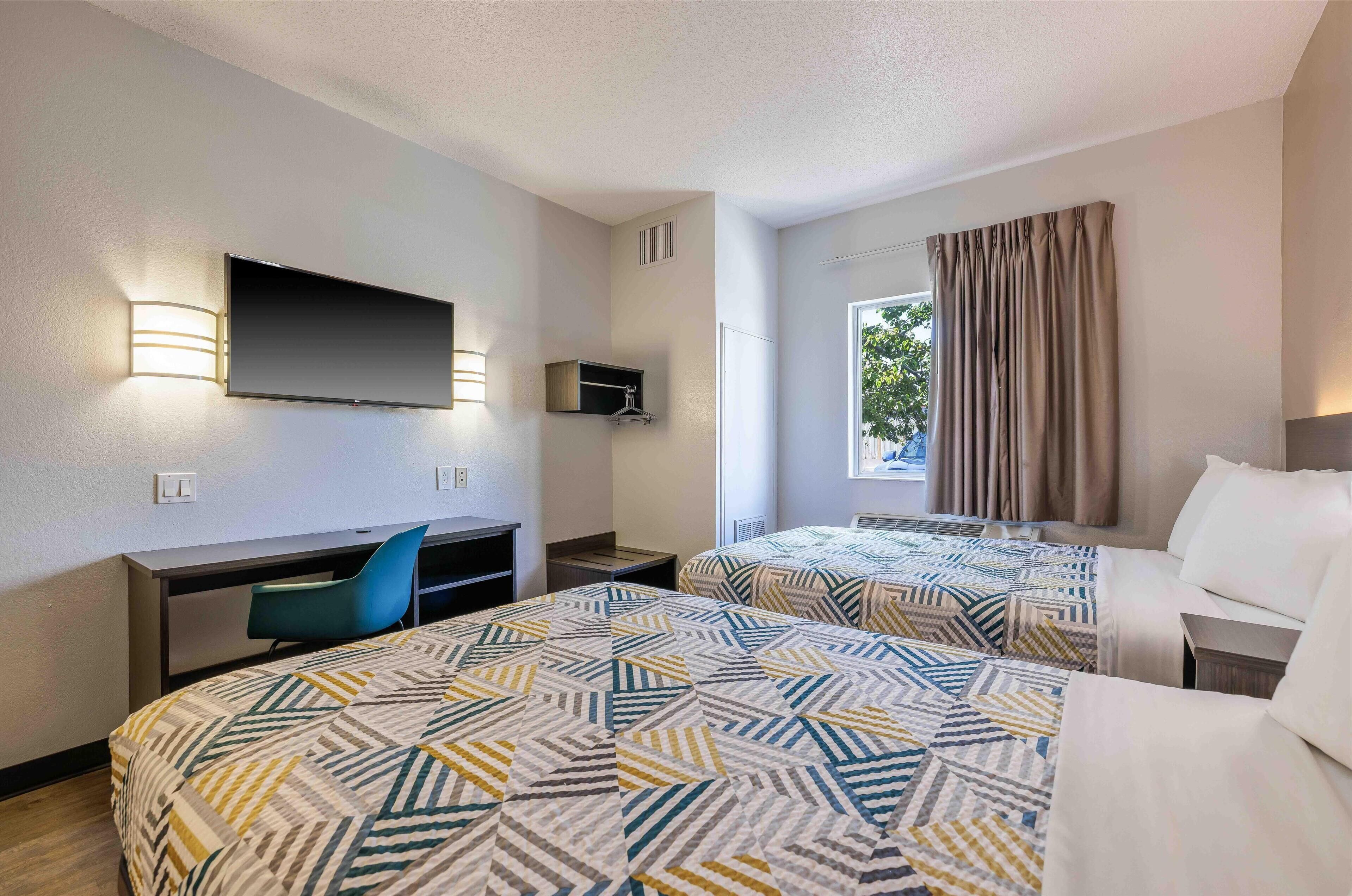 A hotel room with two beds, a desk, a chair, a TV, and a window with curtains.