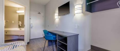 A hotel room with a bed, a desk, a chair, a TV mounted on the wall, and a shower area.