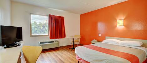 A hotel room with a bed, a desk with a television, a chair, a window with curtains, and a wall-mounted light.