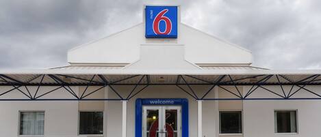 Un motel con un letrero que dice '6', una entrada con la palabra 'Welcome' y una banca afuera.