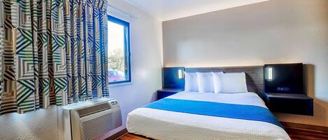 A hotel room with a large bed, a window with patterned curtains, a heater, and a nightstand with a lamp.