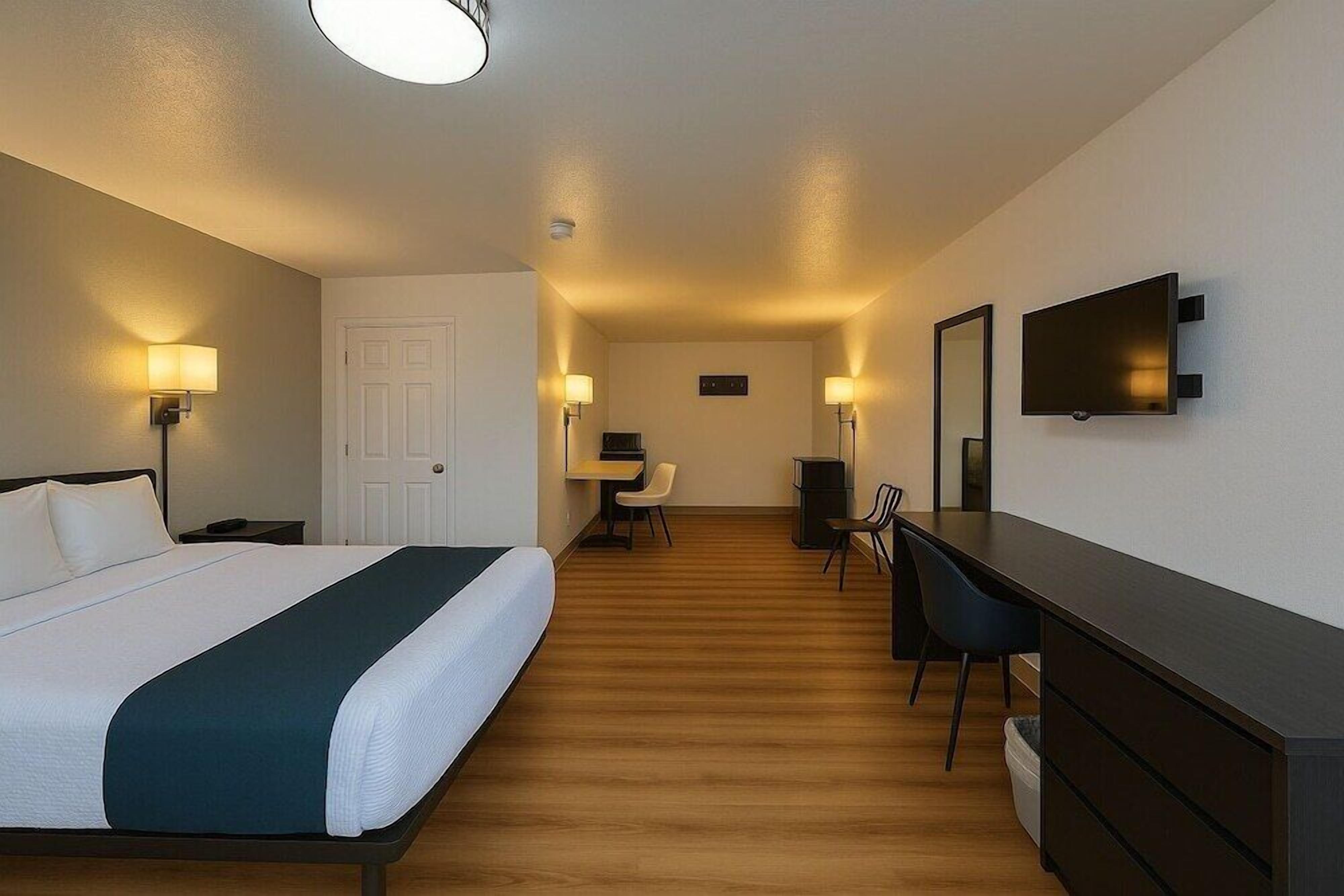 A hotel room with a bed, desk, chair, TV, and a small table with a lamp.