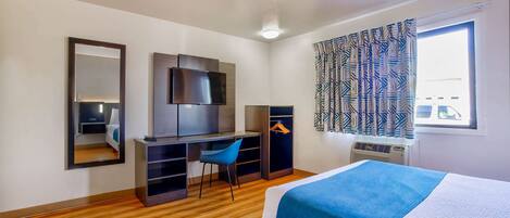 Une chambre d’hôtel comprenant un lit, un bureau, une chaise, une armoire, un téléviseur et une fenêtre avec des rideaux.