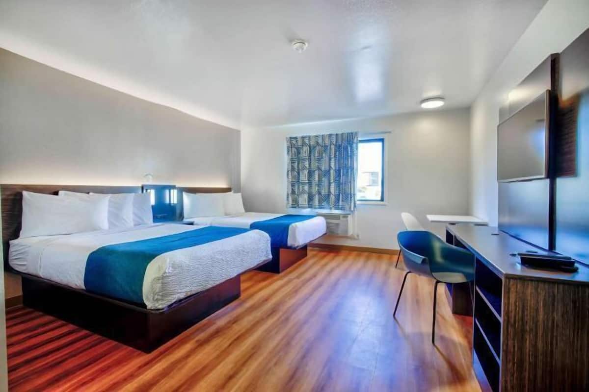 Habitación de hotel con dos camas, un escritorio, una silla, un televisor y una ventana con cortinas estampadas.
